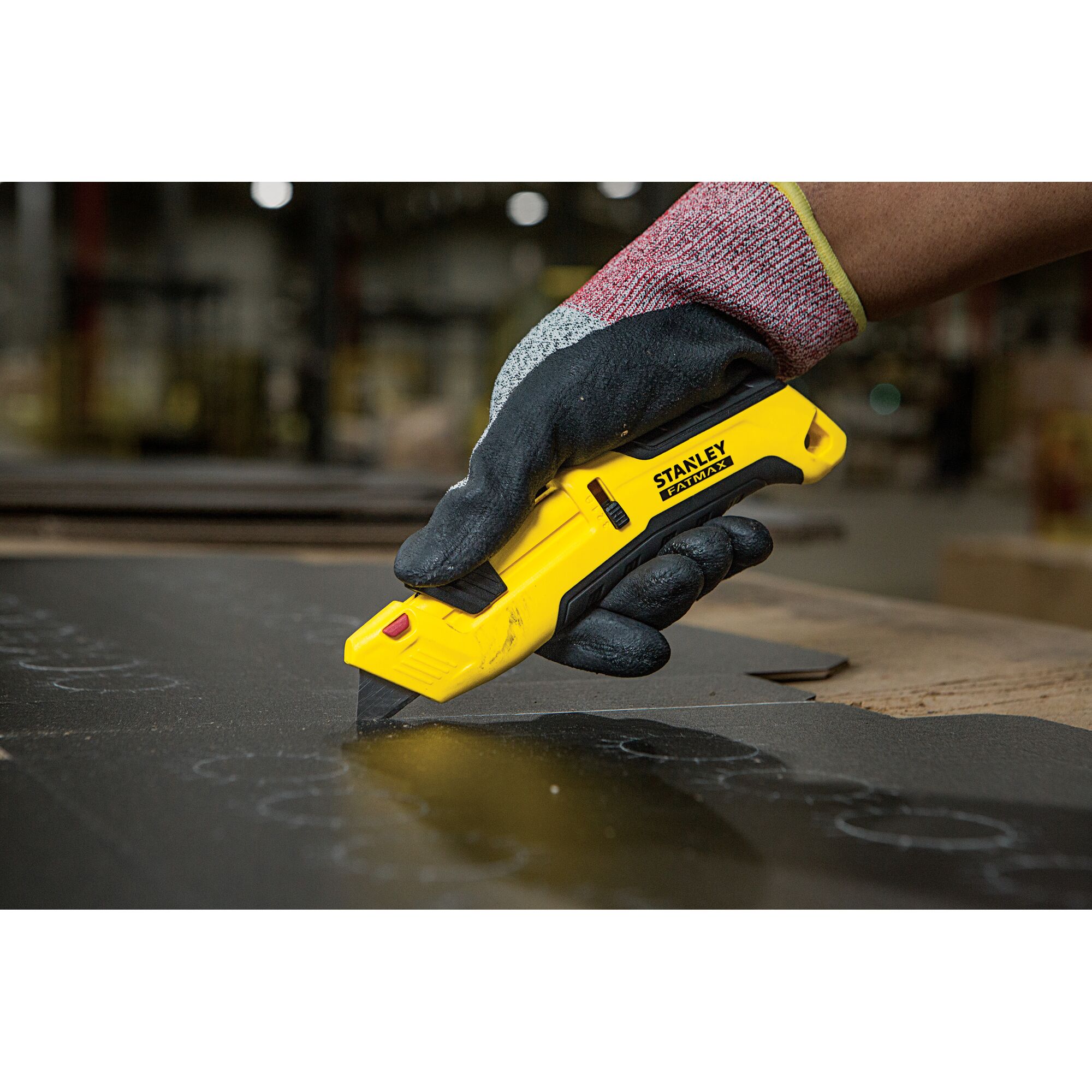Photo of STANLEY FMHT10365-0 utility knife in a gloved hand cutting black material, indoor workshop setting.