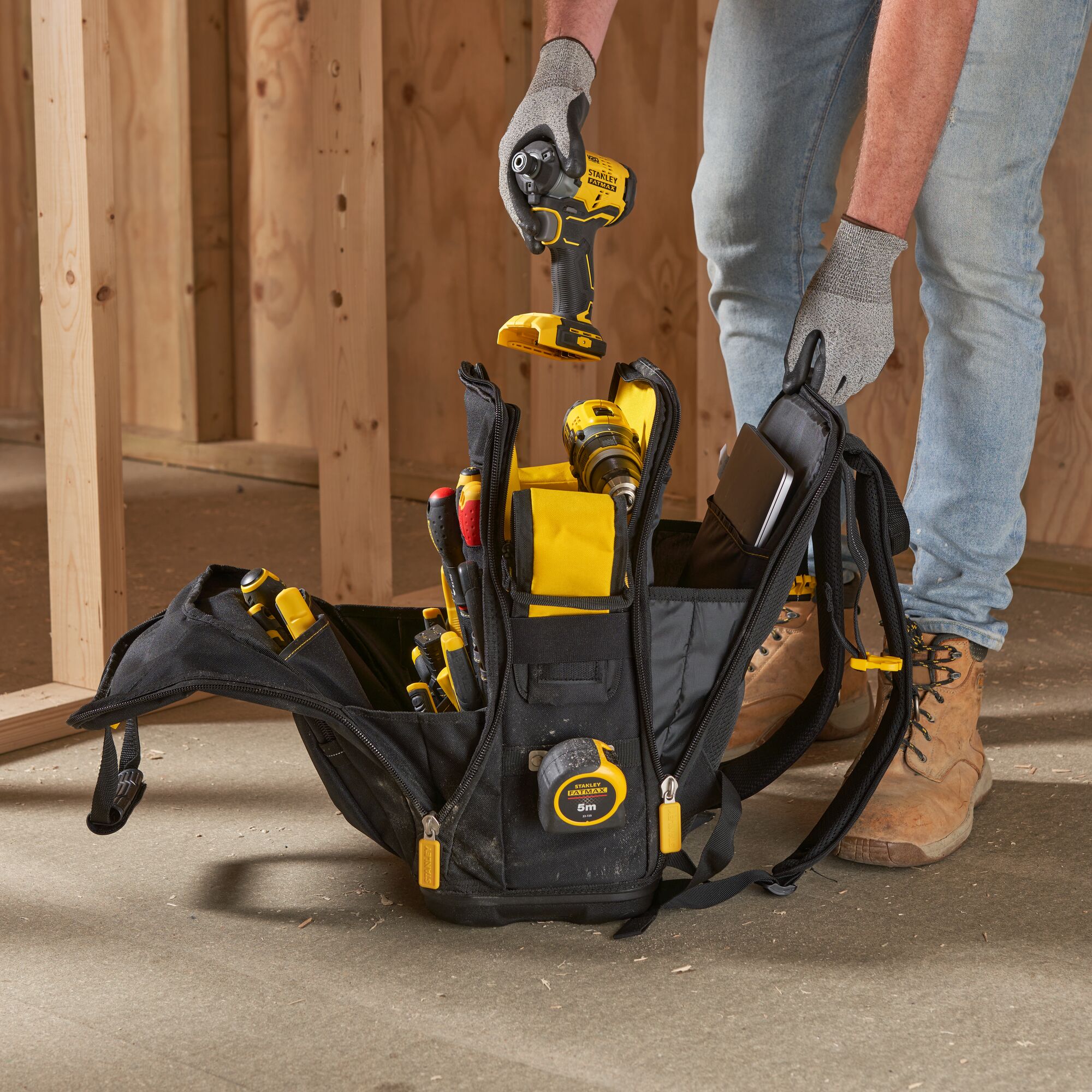 STANLEY FMST1-80144 tool backpack with yellow and black tools on job site floor, worker's hands visible.