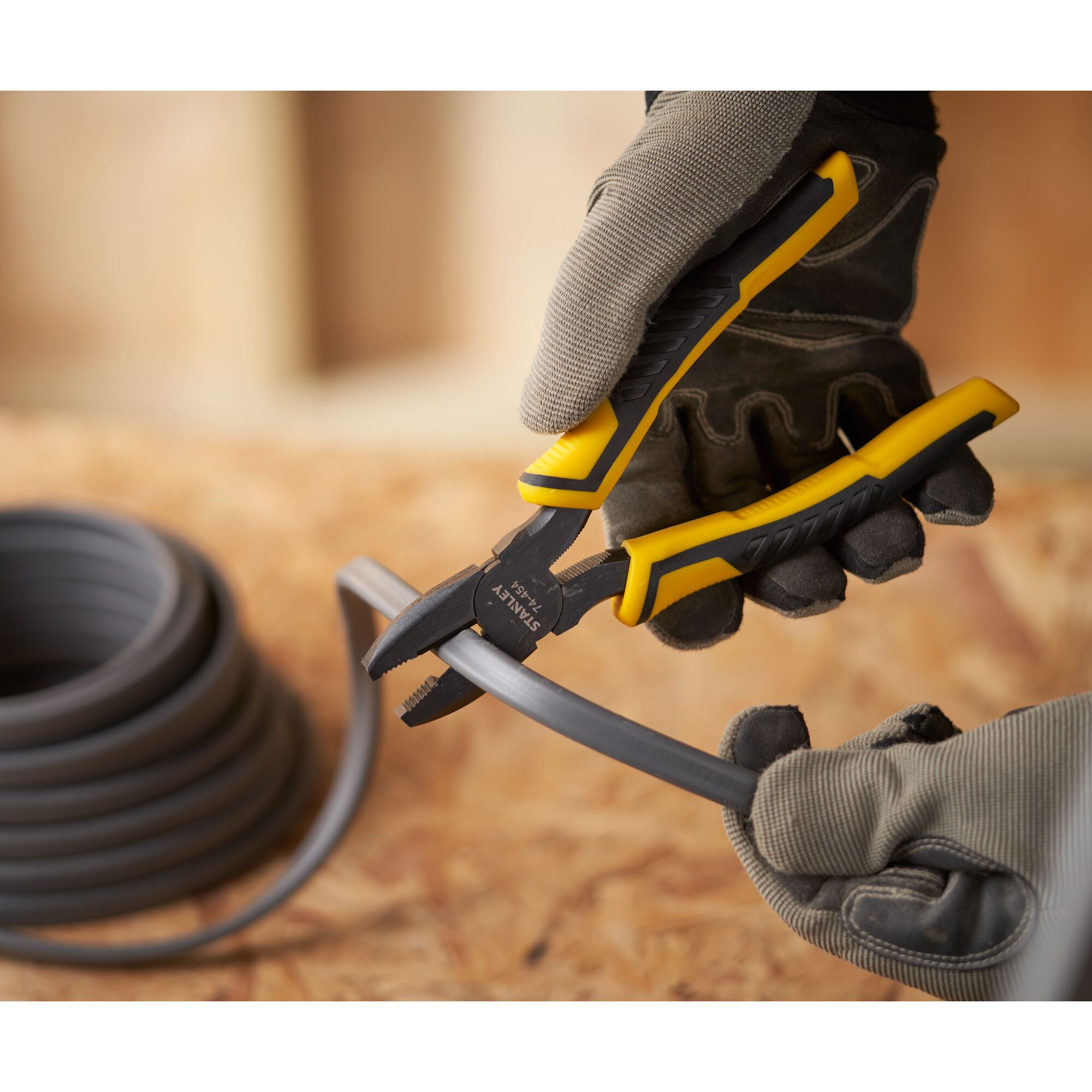 STANLEY STHT0-74456 pliers with yellow and black handle cutting black cable, worn by gloved hand, close-up.