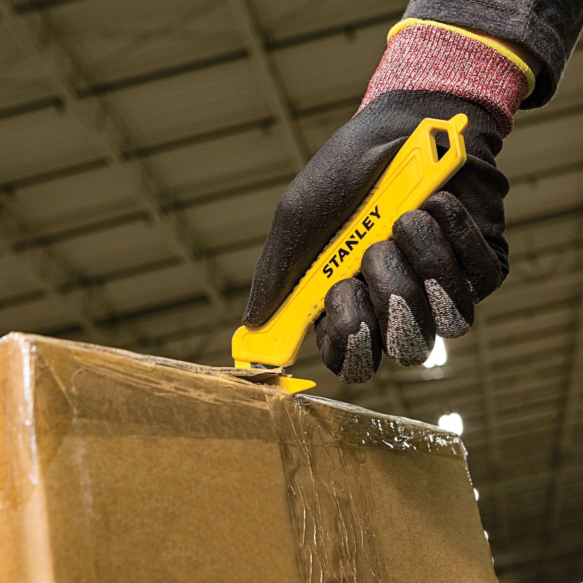STANLEY STHT10355 yellow safety utility knife cutting tape on a cardboard box, held by a gloved hand.