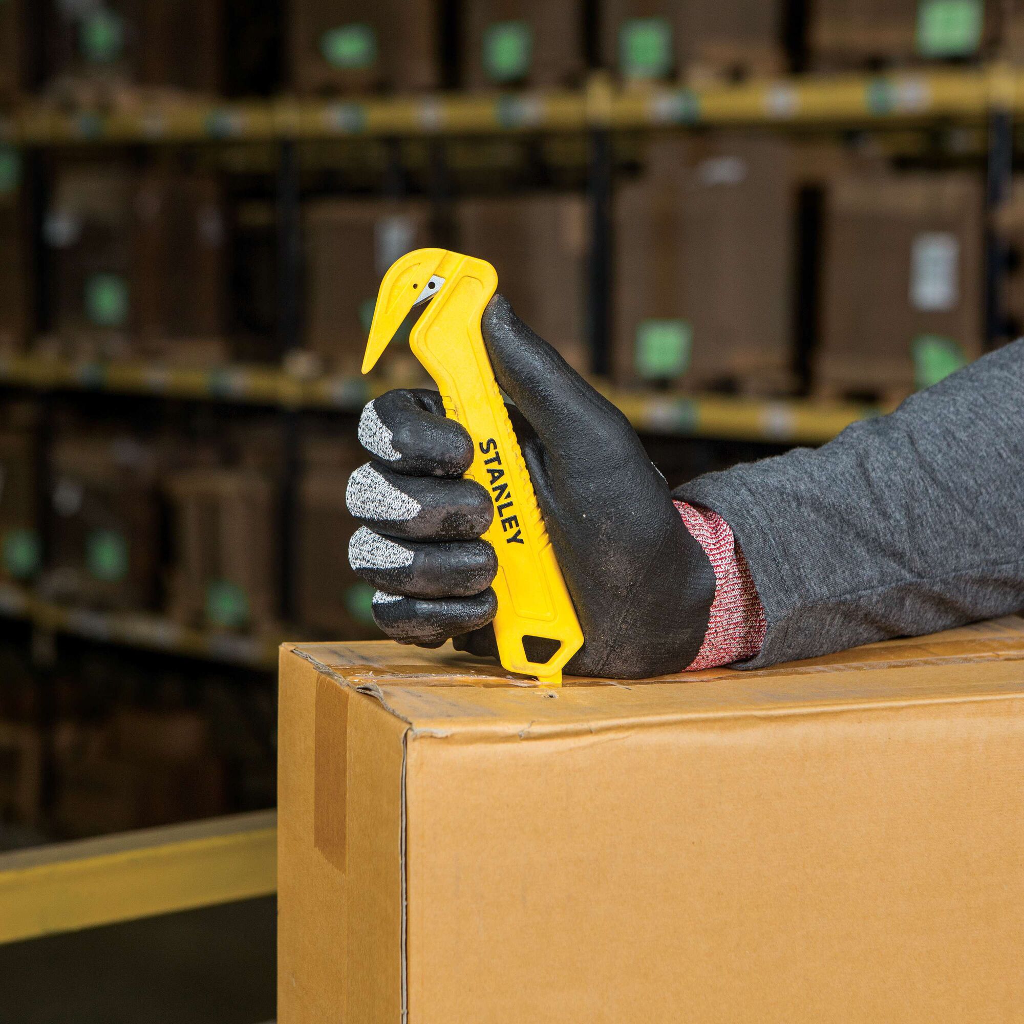 Photo of STANLEY STHT10355 yellow safety box cutter held in gloved hand cutting cardboard box in warehouse.