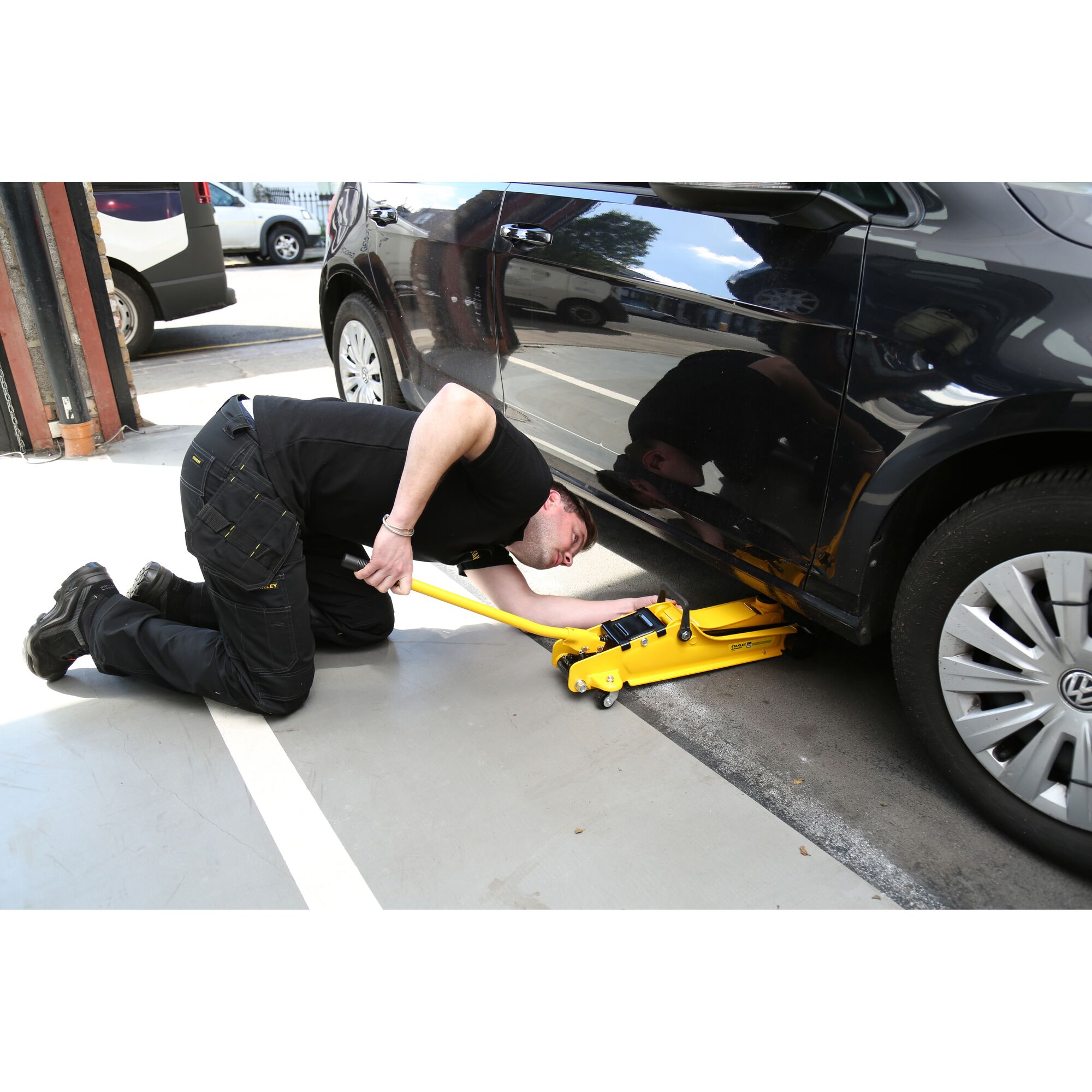 STANLEY STMT81251-1 yellow jack lifting a black car, technician kneeling beside it in workshop.